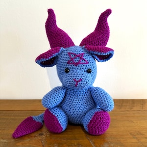 May include: A crocheted plush toy featuring a light blue body with dark purple horns, ears, feet, and a pointed tail. The creature has black safety eyes, a stitched pink nose, and a dark purple pentagram embroidered on its forehead.