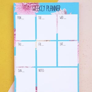 May include: A weekly planner with a blue background and pink floral design. The planner has spaces for each day of the week, Monday through Sunday, and a notes section.