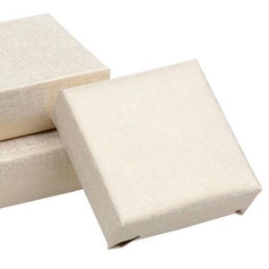 May include: Three small, square, cream-coloured gift boxes with a textured, linen-like appearance. The boxes are stacked, with one slightly open, revealing a glimpse of the interior. The boxes are simple and elegant.