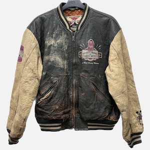 May include: A black and tan leather varsity jacket with a Mickey Mouse Trader Mickey logo on the back. The jacket has a zipper closure and a ribbed collar, cuffs, and waistband.