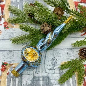 May include: A light blue and gold Christmas tree topper with a spiral design. The topper has a gold glitter base and tip, and a decorative round center. The topper is surrounded by pine needles and pine cones.