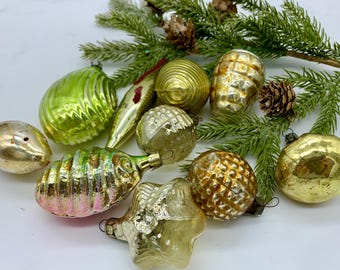 May include: A collection of vintage Christmas ornaments in various shapes and colors, including gold, green, and silver. Some ornaments have textured surfaces, and others are smooth. A sprig of evergreen and pinecones are also present.