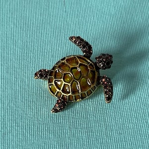 May include: A decorative turtle brooch with a gold-toned frame and a green and yellow shell. The turtle's head, feet, and flippers are embellished with dark red crystals. The brooch is set against a light blue background.