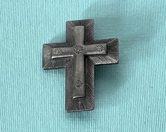 May include: A dark gray metal cross pendant featuring a layered design with a textured, sunburst-style finish on the outer edges. The central vertical and horizontal bars are adorned with small, engraved circular floral motifs at each end.