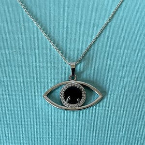 May include: A silver-toned necklace featuring an eye-shaped pendant. The pendant has a central round black stone surrounded by a halo of small clear stones, all set within a polished silver-toned metal frame suspended from a delicate chain.
