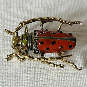 May include: A gold-toned beetle brooch featuring a textured metallic body with long, jointed legs. The insect has a vibrant red wing casing decorated with small black spots, a grey midsection, and two bright green accents near the head.
