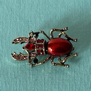 May include: A red and gold beetle brooch with rhinestones. The beetle has a shiny red body and gold legs and antennae. The beetle is decorated with clear and pink rhinestones.