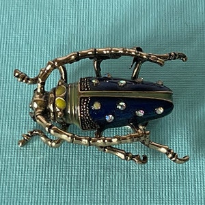 May include: A gold-toned beetle brooch with blue enamel and clear rhinestones. The beetle has yellow accents on its head and a green stripe around its body.