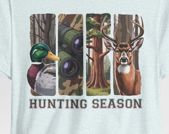May include: Light blue t-shirt featuring a graphic with four vertical panels. The panels depict a duck, binoculars, a tree, and a deer. The words "HUNTING SEASON" are printed in a bold font below the panels. The shirt is made of a soft material.