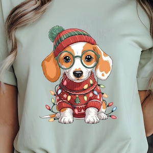 May include: A light green t-shirt featuring a cartoon beagle wearing a red sweater, hat, and glasses, surrounded by Christmas lights. The beagle has orange and white fur. The shirt is made of a soft material.
