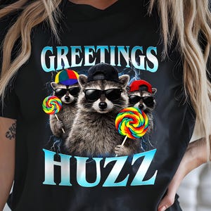 Op de afbeelding: Zwart T-shirt met een afbeelding van drie wasberen met een zonnebril en lolly's. De tekst "GREETINGS HUZZ" staat boven en onder de wasberen in een blauw verlooplettertype. De wasberen dragen hoeden in verschillende kleuren.
