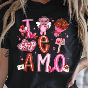 Op de afbeelding: Zwart T-shirt met een Valentijnsdagontwerp. Het ontwerp bevat de woorden "Te Amo" in rood en roze, samen met illustraties van een teddybeer, harten, rozen en enveloppen. Het shirt is een crewneck met korte mouwen.
