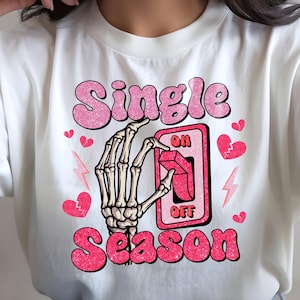 May include: White t-shirt with a pink graphic that reads "Single Season." The design includes a skeleton hand flipping an "on/off" switch, hearts, and lightning bolts. The text is in a bubble letter style.