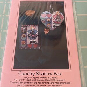 May include: A pink and white pattern sheet for a country shadow box craft project. The pattern includes a rag doll, apples, flowers, and hearts. The pattern is 9 1/2 inches by 11 inches. The text on the pattern reads "Country Shadow Box" and "#9".