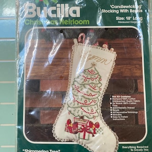 May include: A Bucilla Christmas Heirloom candlewicking stocking kit in plastic packaging. The kit features an 18-inch long stocking with a white fabric base, a green embroidered Christmas tree, red beads, and gifts. The text reads Shimmering Tree.