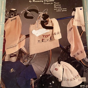 May include: A book titled "Fingertips in Action" by Rosemary Drysdale. The book cover features a bicycle with a white bag that says "TOM" and a white towel that says "MIKE". The book cover also features a blue shirt with a cyclist graphic, a white towel with the letters "RD" and a black helmet.