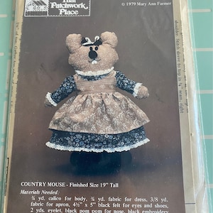 May include: A sewing pattern for a Country Mouse doll, finished size 48 cm tall. The doll is wearing a dress with a floral pattern and a white collar. The pattern includes instructions and material requirements.