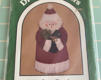 May include: A pattern for a Santa Claus Lane wall or door hanging. The pattern is from Dream Spinners and is #145. The pattern is for a Santa Claus with a red and white suit, a white beard, and a black hat. The pattern is copyrighted 1988 by Great American Quilt Factory, Inc.