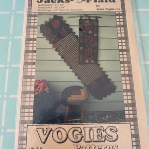 May include: A sewing pattern for a Halloween-themed wall hanging, table runner, and stool cover. The pattern is called "Jacks-Plaid" and is by June Vogitance. The pattern includes instructions for making a 23 cm x 61 cm wall hanging, a 38 cm x 137 cm table runner, and a 30 cm round stool cover.