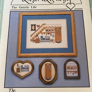 May include: A cross stitch pattern booklet titled "HeartStrings: The Gentle Life" featuring a home with a staircase and the text "Home Hearts Happiness". The booklet also includes three smaller cross stitch patterns: a heart-shaped pattern with a kitchen scene, a rectangular pattern with a door, and a rectangular pattern with a welcome sign.