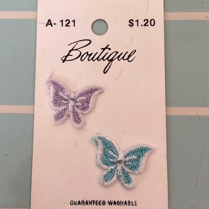 May include: Two iron-on embroidered butterfly patches on a white card. The butterflies are purple and blue with white outlines. The card has the text "Boutique", "A-121", "$1.20", "Guaranteed Washable", "Sz", and "1 On".