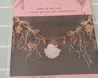 May include: A pink and white paper pattern for a Christmas garland with a Santa Claus ornament. The pattern is titled "Bits & Pieces by Joan" and includes the text "Santa on the Line, A Santa Garland plus ornament/pin".