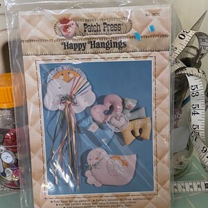 May include: A craft kit for making three wall hangings. The kit includes a pattern with instructions for making a cloud with a rainbow, a bird, and a heart. The kit is called "Happy Hangings" and is from Patch Press.