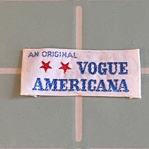 May include: A rectangular white label with the text "AN ORIGINAL" above "VOGUE AMERICANA". Two red stars are between the words. The text is in blue, and the stars are red. The label is on a light green surface.