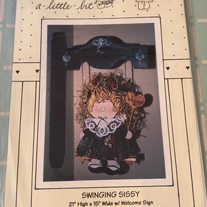 May include: A craft pattern for a wooden doll hanging from a swing. The doll is wearing a black dress with a white lace collar and has a straw-like hairdo. The doll is 53 cm high and 38 cm wide. The pattern is called "Swinging Sissy" and includes a welcome sign.