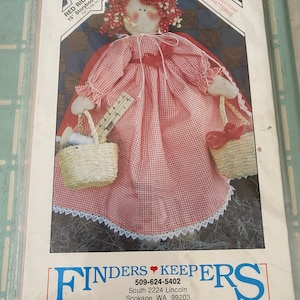May include: A sewing pattern for a 15-inch Red Ridin' Hood Storybook Folk Doll. The pattern includes complete instructions and full-size patterns. The doll is wearing a red and white gingham dress with a red cape and carrying two baskets.