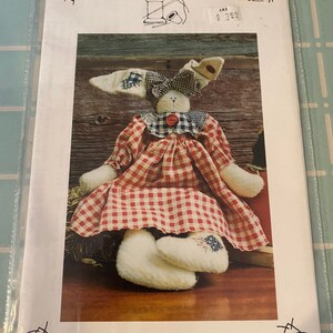 May include: A vintage sewing pattern for a red and white gingham bunny doll. The pattern is titled "A Stitch In Time" and is number 102. The pattern is from the company "Patches".