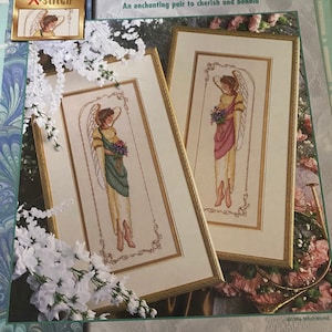 May include: Elegant Angels cross stitch pattern featuring two angels with wings, one in a green dress and the other in a pink dress, both holding bouquets of flowers. The pattern is framed and measures 11 inches by 20 inches.