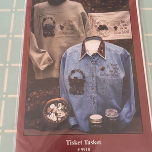 May include: A pattern for a cross stitch project called "Tisket Tasket" featuring a basket of buttons. The pattern is by Olde World Reflections and is number 9918. The pattern shows a sweatshirt, a shirt, and a small basket of buttons.