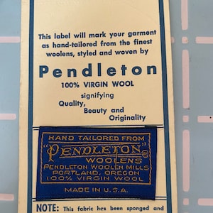 May include: A vintage Pendleton label, cream-colored with blue text and accents. The label reads "Pendleton 100% Virgin Wool" and includes a smaller blue tag with gold lettering. The label also states "Made in U.S.A."