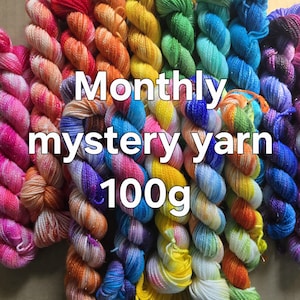 May include: A collection of colorful yarn skeins in various shades, including pink, orange, yellow, green, blue, and purple. The text "Monthly mystery yarn 100g" is overlaid on the image. The yarn is arranged in vertical rows.