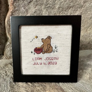 Puede incluir: Un bordado enmarcado con un oso de dibujos animados sosteniendo una olla de miel. El bordado está sobre un fondo blanco con texto rojo que dice "Liam Joseph 6 de julio de 2023".