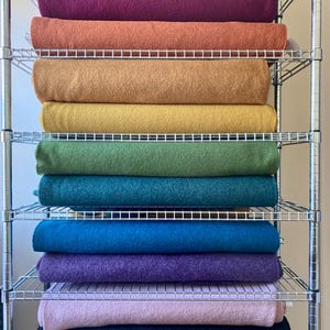 May include: A metal shelving unit displays a collection of rolled blankets in various colors. The blankets range from pink to deep blue, with shades of orange, yellow, and green. The blankets are neatly stacked on the wire shelves, creating a visually appealing display.