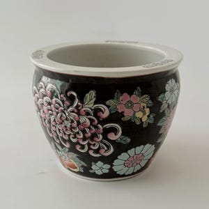 May include: A round ceramic bowl with a black exterior featuring a colorful floral pattern of pink, white, and light blue chrysanthemums and green leaves. The interior and the top rim are white, with small decorative symbols along the upper edge.