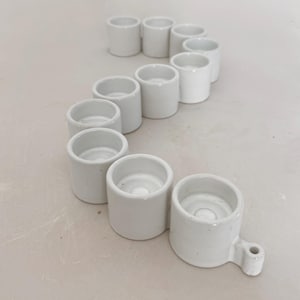May include: A collection of small, white ceramic candle holders arranged in a curved line. Each holder is cylindrical with a recessed top for a candle. One holder has a small handle. The holders are on a neutral-toned surface.