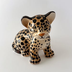 May include: A ceramic figurine of a seated leopard cub with a glossy finish. The cub features a tan base coat covered in dark brown to black spots, with painted green eyes, a small brown nose, and large ears. The sculpture is depicted in a sitting pose.