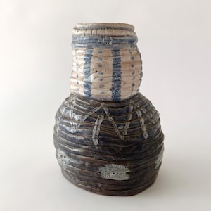 May include: A handcrafted ceramic vase with a textured surface. The upper section features blue and white stripes, while the lower portion is a darker, textured brown. The vase has a unique, organic shape, suitable for home decor.