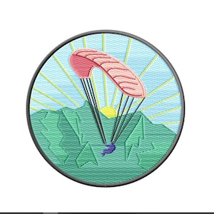Può includere: Una toppa rotonda ricamata con un parapendio rosa e bianco che vola sopra montagne verdi con un sole giallo sullo sfondo.