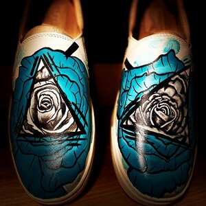 May include: A pair of white canvas shoes with a hand-painted design. The design features a blue rose with a black triangle and eye inside.