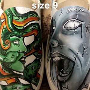 May include: A pair of white canvas slip-on shoes with hand-painted designs. One shoe features a green snake with orange accents, and the other shoe features a blue and white face with a cracked texture. The shoes are size 9 and say "For sale Ugg's-Womens size 9" on the inside.