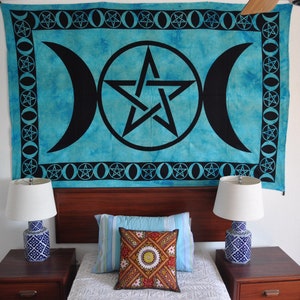 Könnte beinhalten: Ein blauer und schwarzer Wandteppich mit einem Pentagramm und einem Dreifachmond-Symbol hängt über einem Bett mit weißen Bettwäsche und zwei Nachttischen mit Lampen.