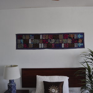 May include: A colorful patchwork tapestry with a purple border hangs on a white wall above a wooden headboard with a white and brown pillow. A green plant is in the corner of the room.