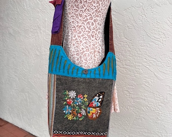 May include: A colorful patchwork shoulder bag with a butterfly and floral embroidery on a gray fabric panel. The bag has a blue, green, and brown strap and a maroon bottom.