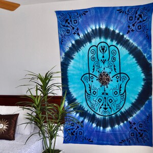 May include: A blue and black tie-dye tapestry with a large, black hamsa hand design in the center. The hamsa hand has intricate details and a small, black flower in the center.