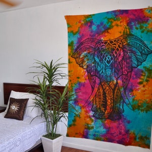 May include: A colorful tie-dye tapestry featuring a black elephant mandala design. The tapestry is hung on a white wall in a bedroom setting.