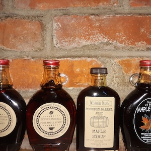 May include: Four glass bottles of maple syrup with red and brown lids. The bottles are labeled with different flavors: Cinnamon Infused Pure Maple Syrup, Coffee Infused Pure Maple Syrup, Bourbon Barrel Aged Maple Syrup, and Pure Ohio Maple Syrup. The bottles are arranged in a row on a brick wall background.